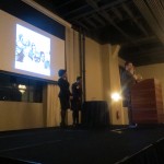 Emcee Latasha Fox & Executive Director Rhonda Gray watch as awardee Corey Smale of Strange Donuts accepts his award